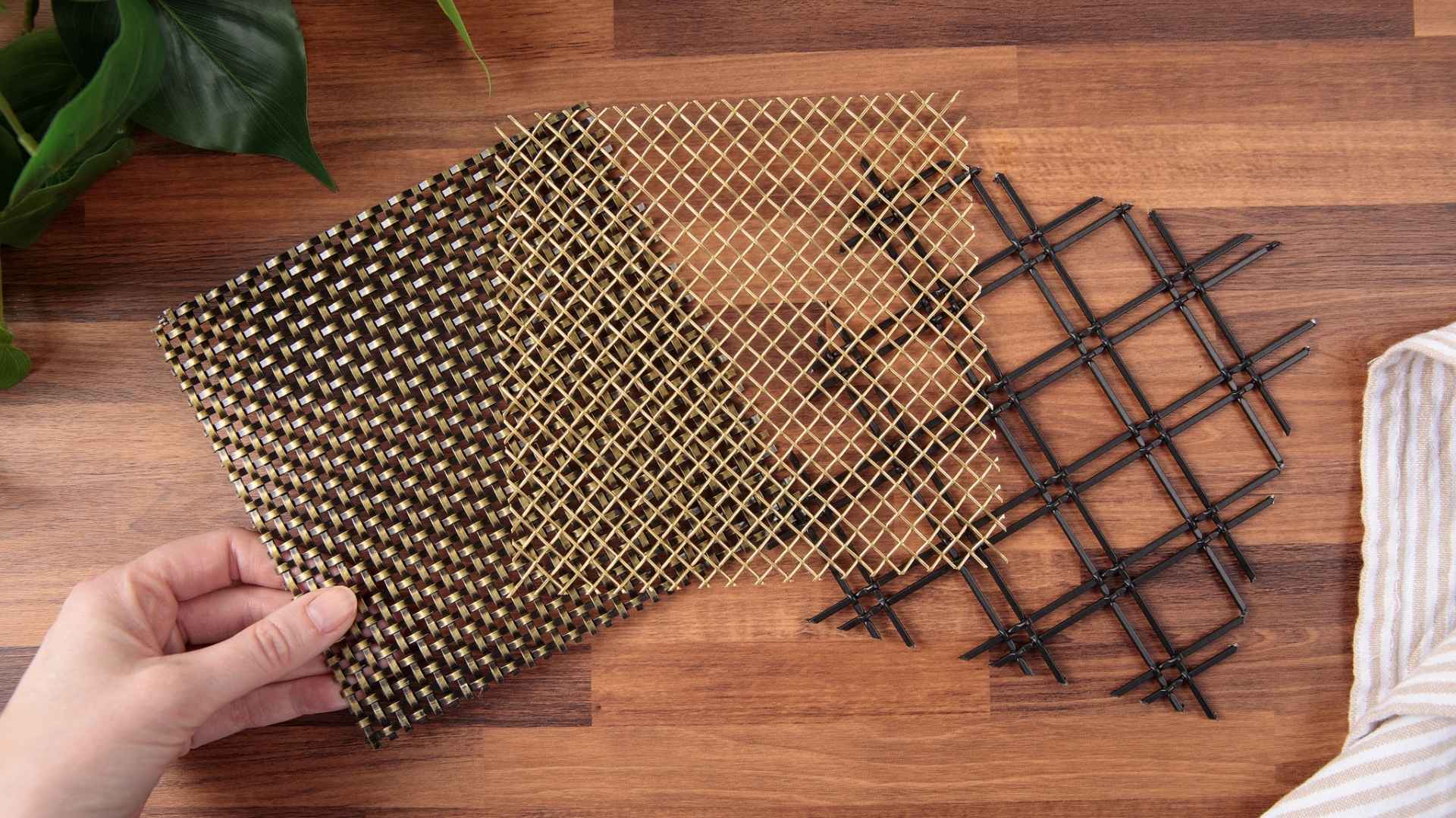 Three decorative wire mesh samples in bronze weave, open brass grid, and black grid patterns displayed on a wooden surface.