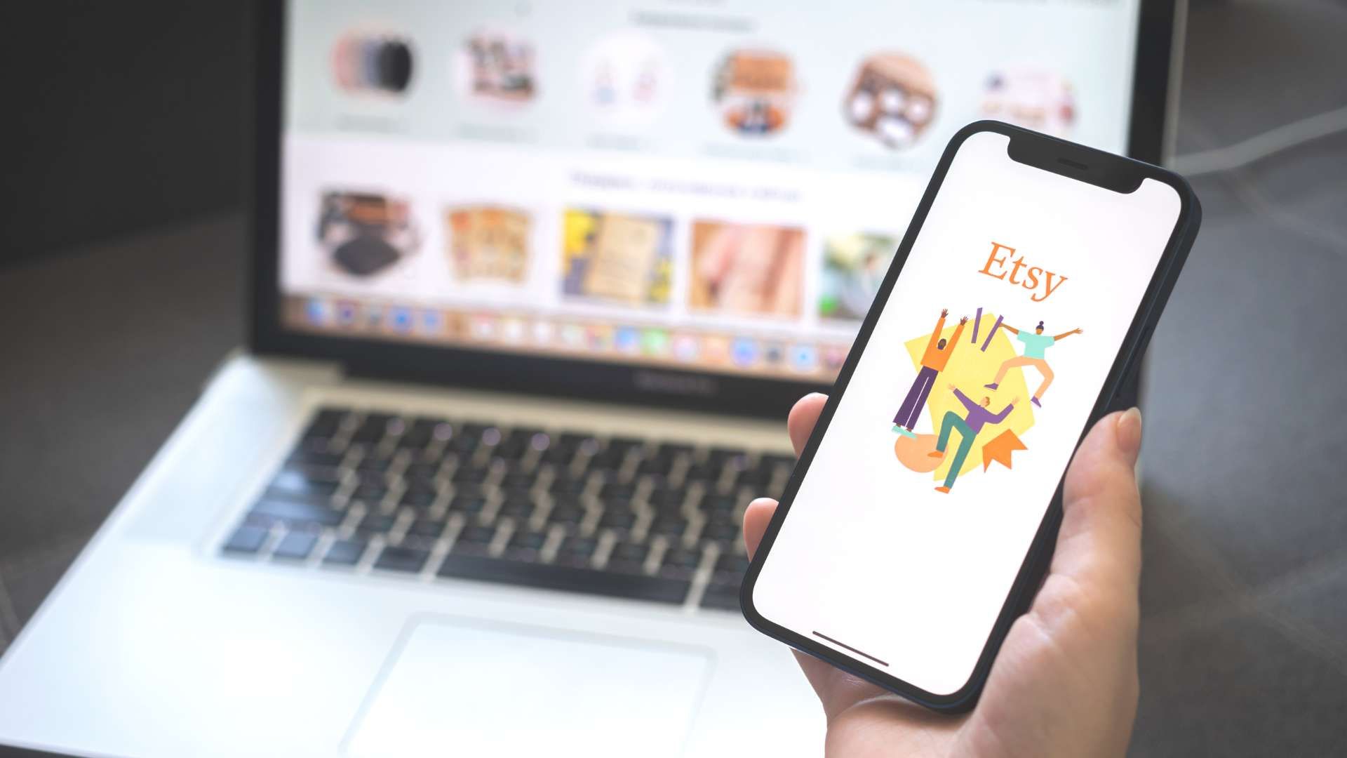 A woman holds a smartphone showing the Etsy website and holds it in front of her laptop which shows items for sale on Etsy.