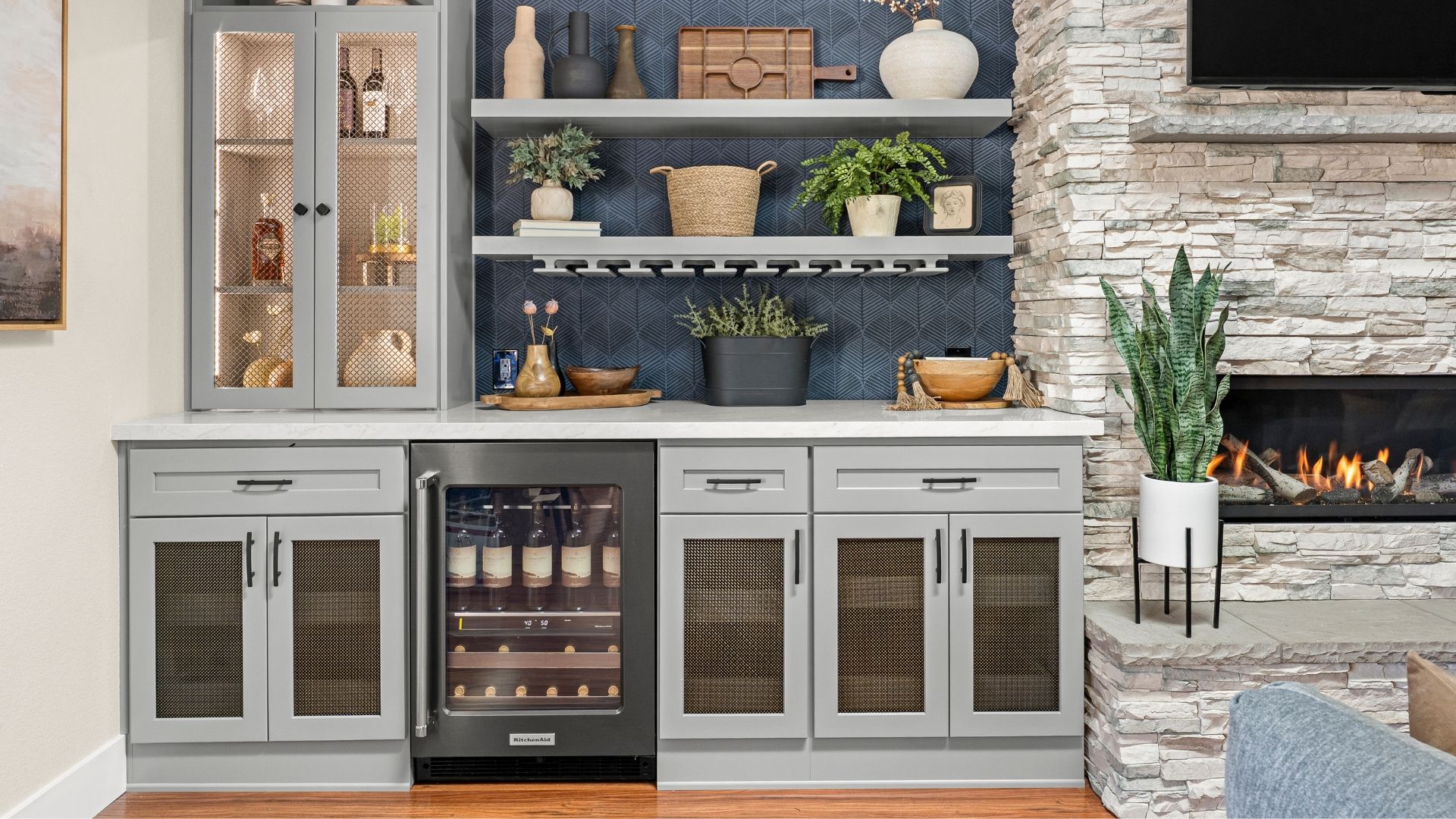 Wet bar with grey cabinets featuring wire mesh cabinet inserts, blue walls, wine fridge, plants, and stone fireplace.