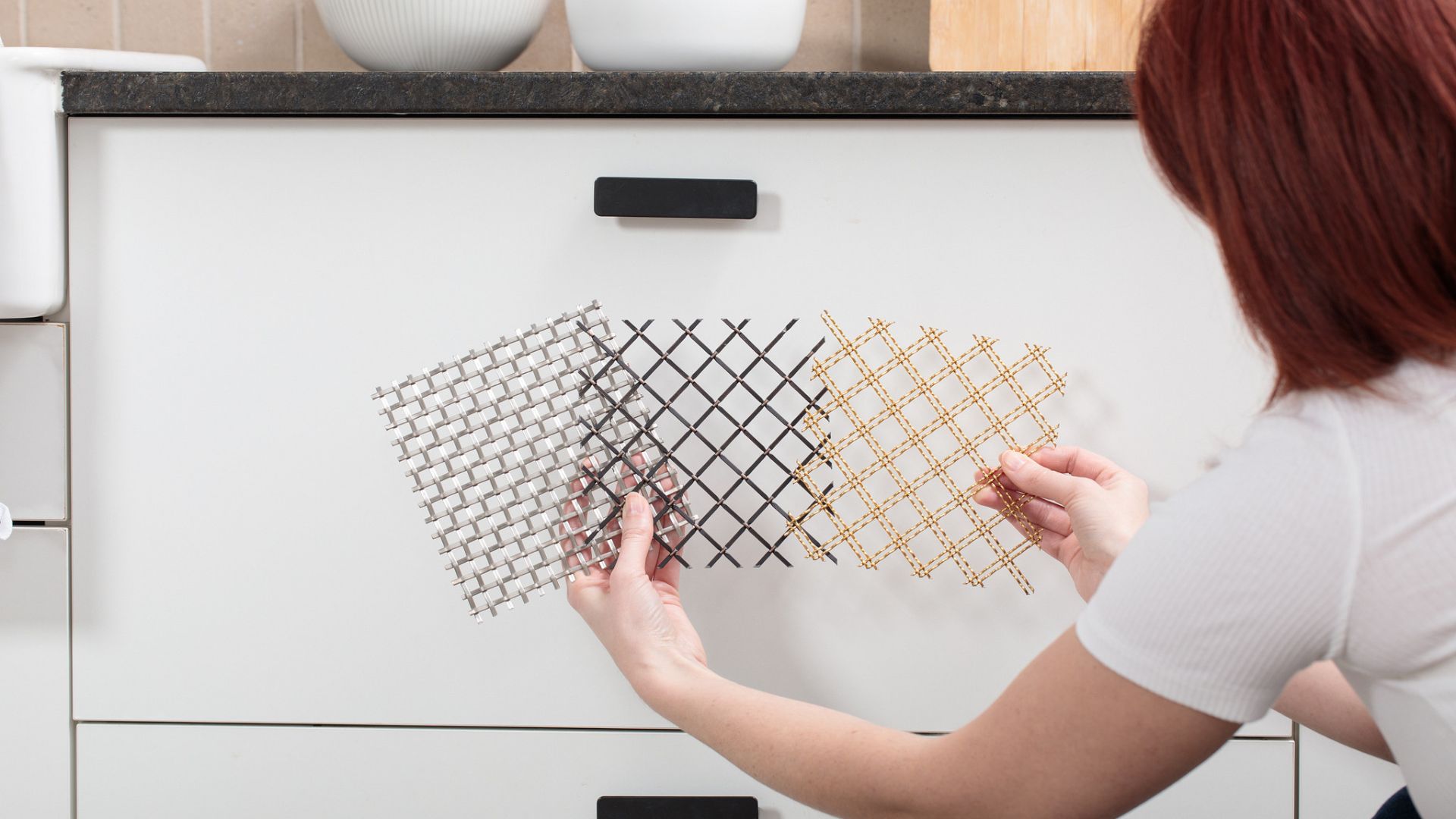 Person holding three overlapping decorative wire mesh samples in silver, black, and brass finishes against a white cabinet.