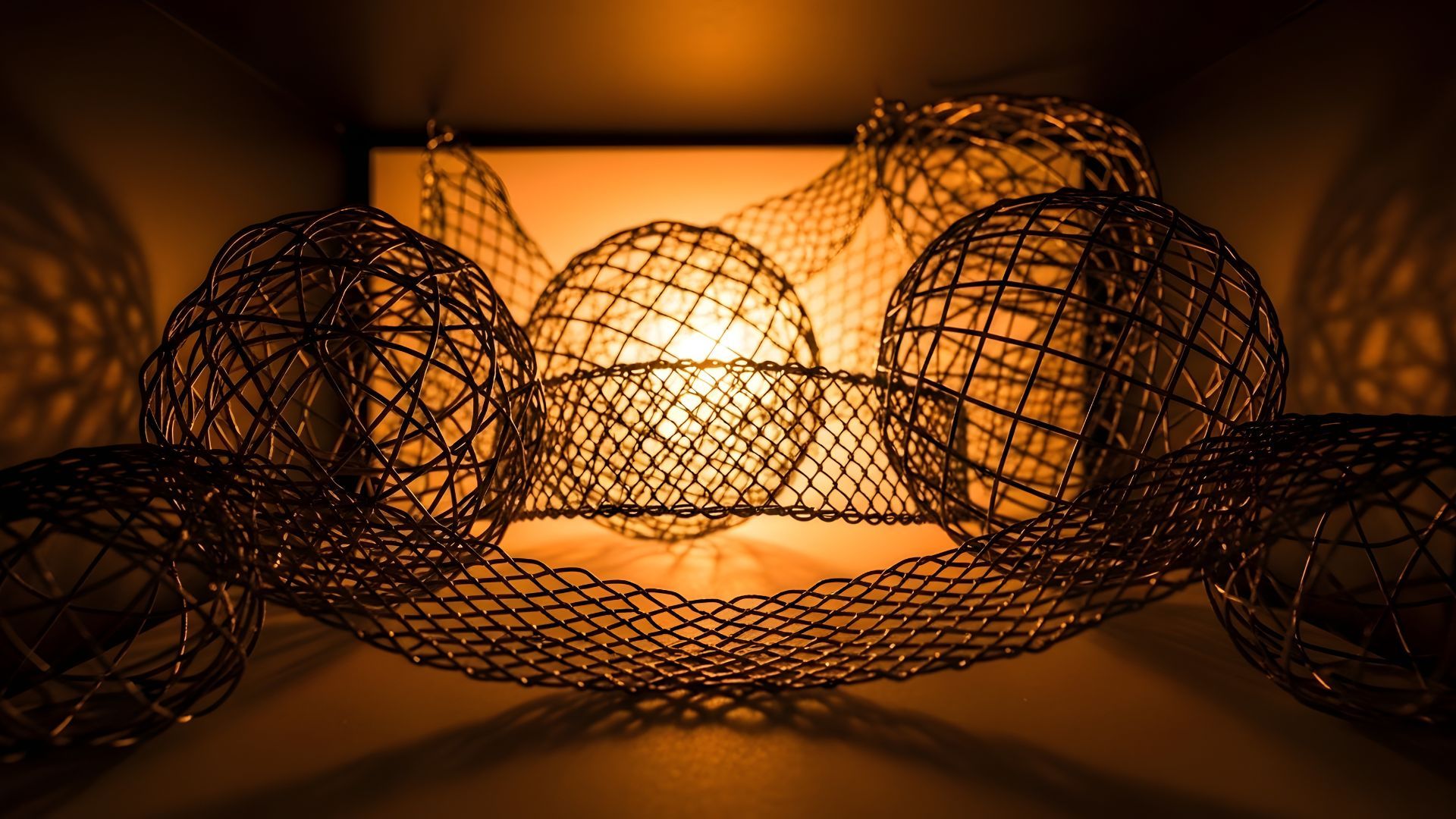 Decorative wire mesh spheres connected by woven metal netting, backlit by a warm orange glow casting intricate shadow patterns.