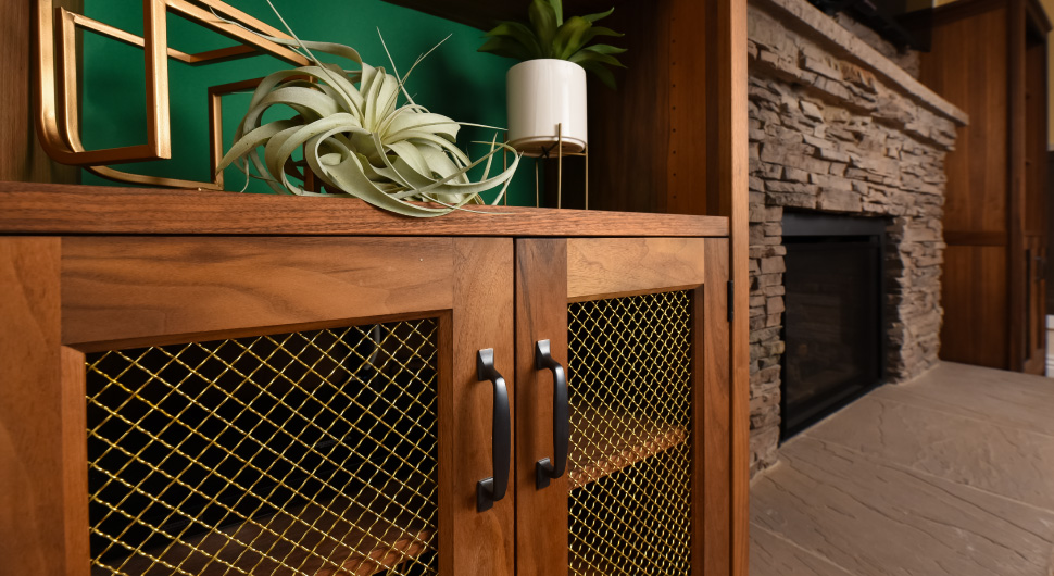 A large wood cabinet in a living room has brass wire mesh inserts and is next to a fireplace with a slate surround.