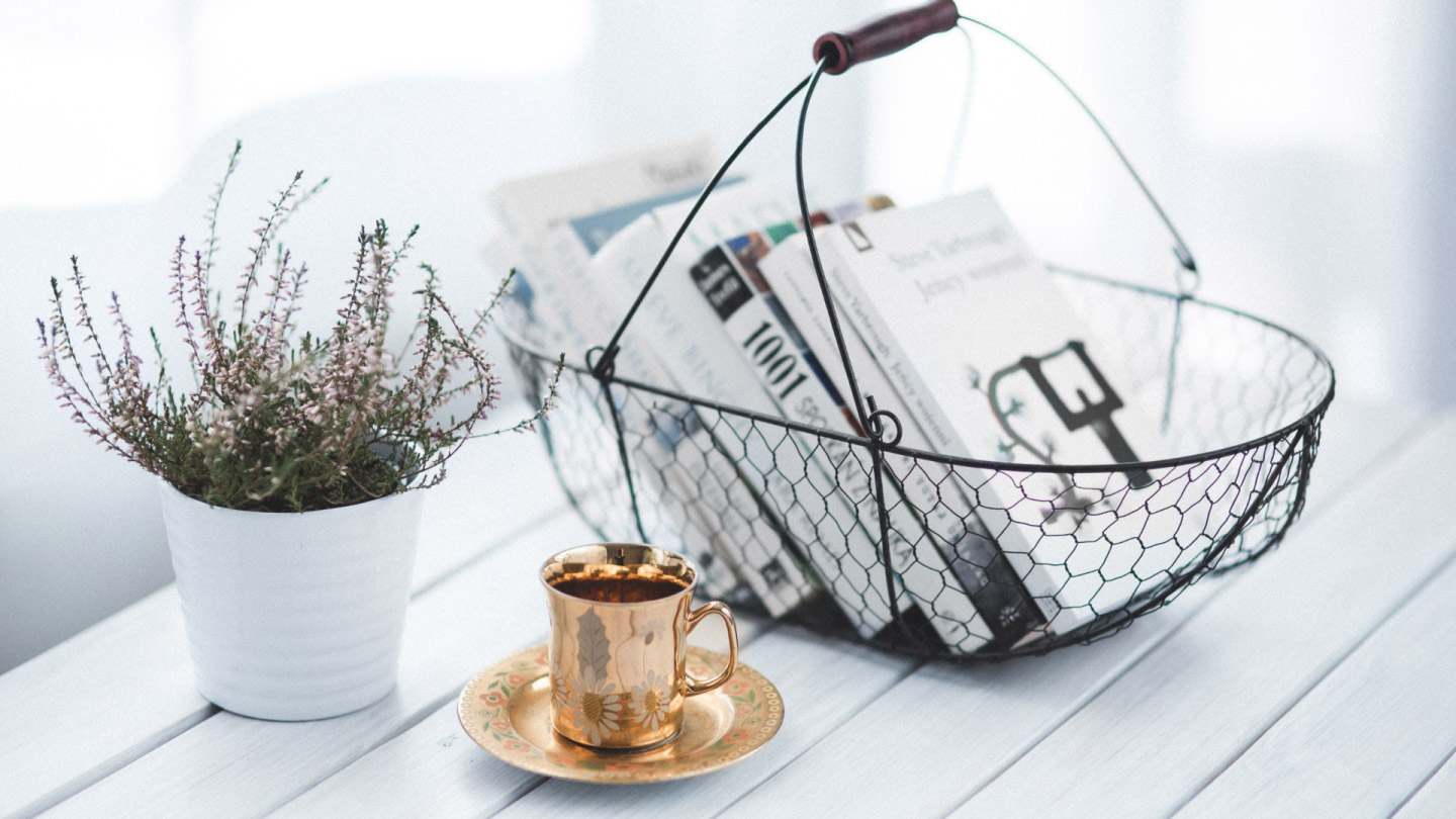Wire mesh basket, plant with white pot, and brass tea cup are placed on a white wood table.