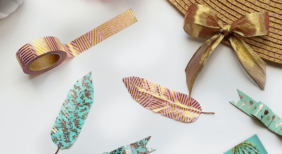 On a desk are a variety of craft materials and projects, including a roll of gold and fuchsia metallic ribbon, handmade paper feathers, and a gold bow made of ribbon.
