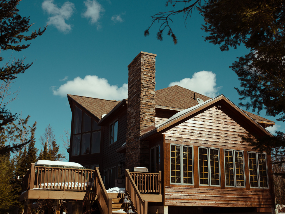 Rustic, lodge-like home in a winter setting with brick chimney, staircase, and large windows.