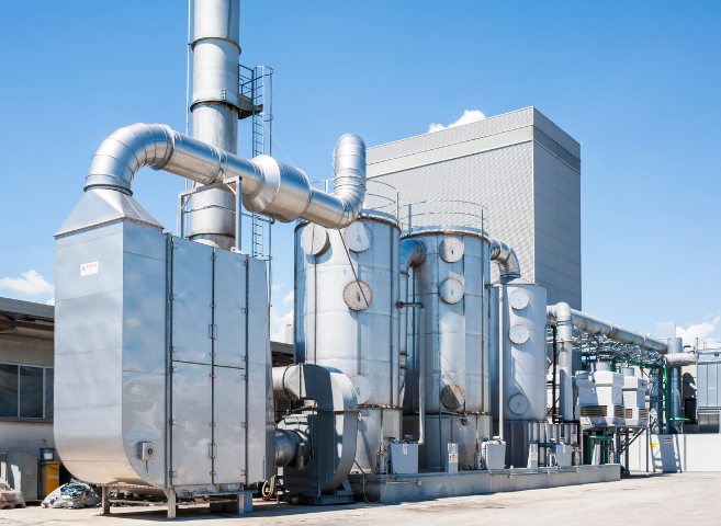 An industrial facility with large metallic pipes, tanks, and machinery set against a bright blue sky with some clouds, likely part of a manufacturing or processing plant.