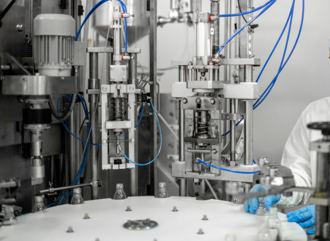 A close-up view of industrial machinery involved in a manufacturing or laboratory process, with blue tubing, metal components, and a worker wearing gloves and a lab coat operating or overseeing the equipment.