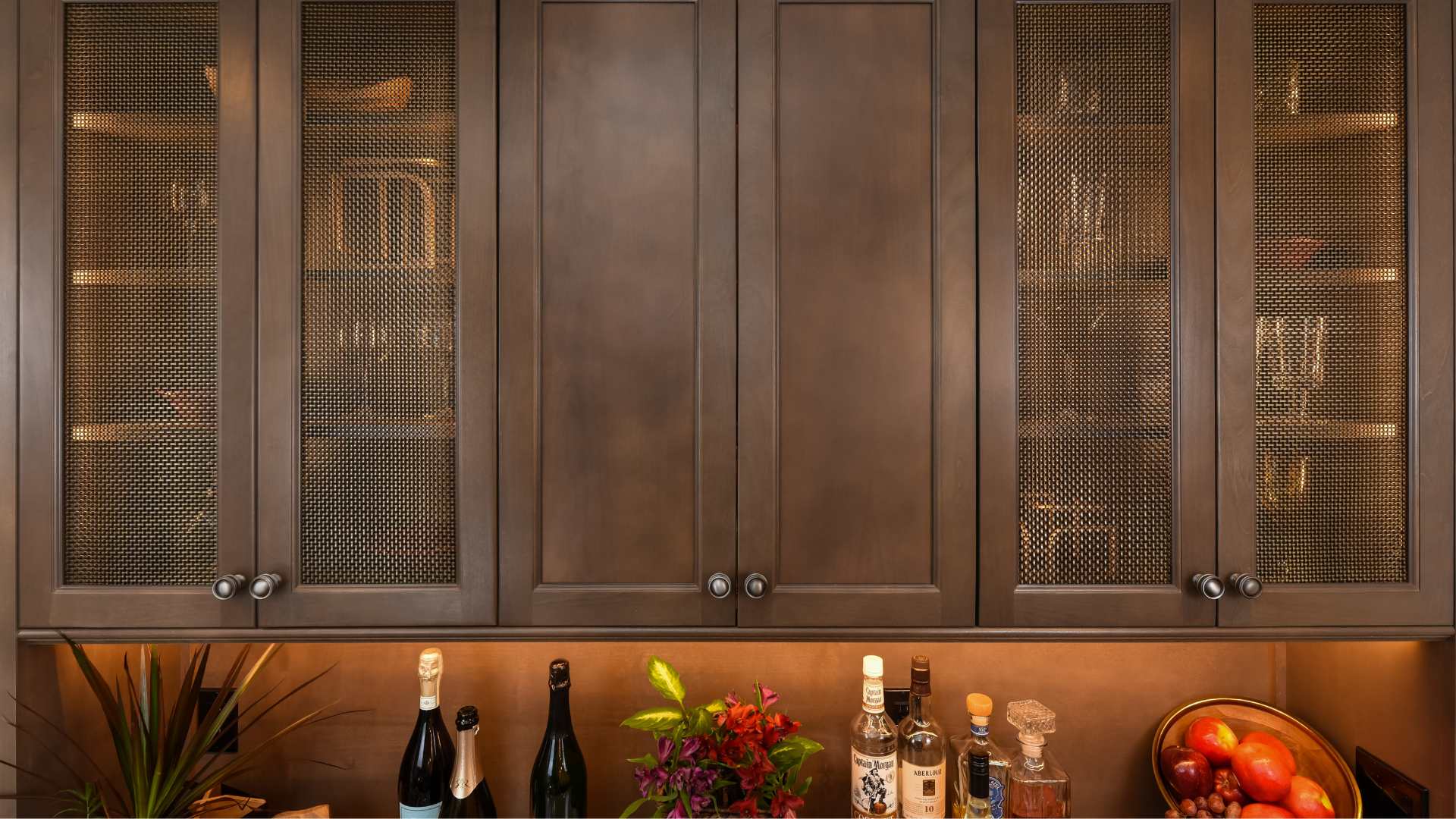 Wet bar that has dark wood cabinets as well as brass cabinet door inserts and under-cabinet lighting.